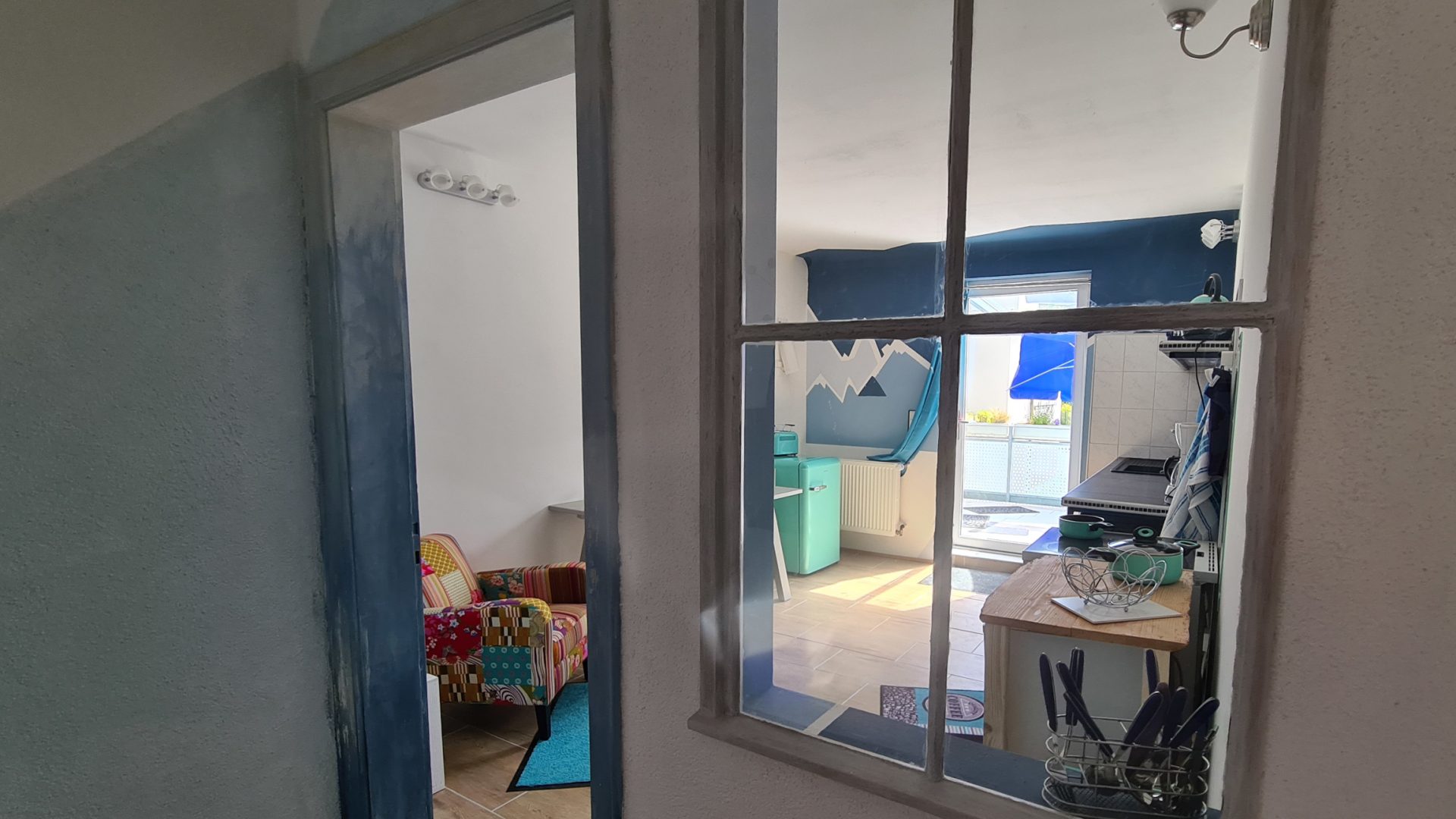 Blick durch ein altes innen eingebautes Sprossenfenster in eine sonnige Wohnküche durch die Glastür sieht man bis zur Terrasse. Einrichtung ist türkis-blau-petrol mit einem bunten Sessel.